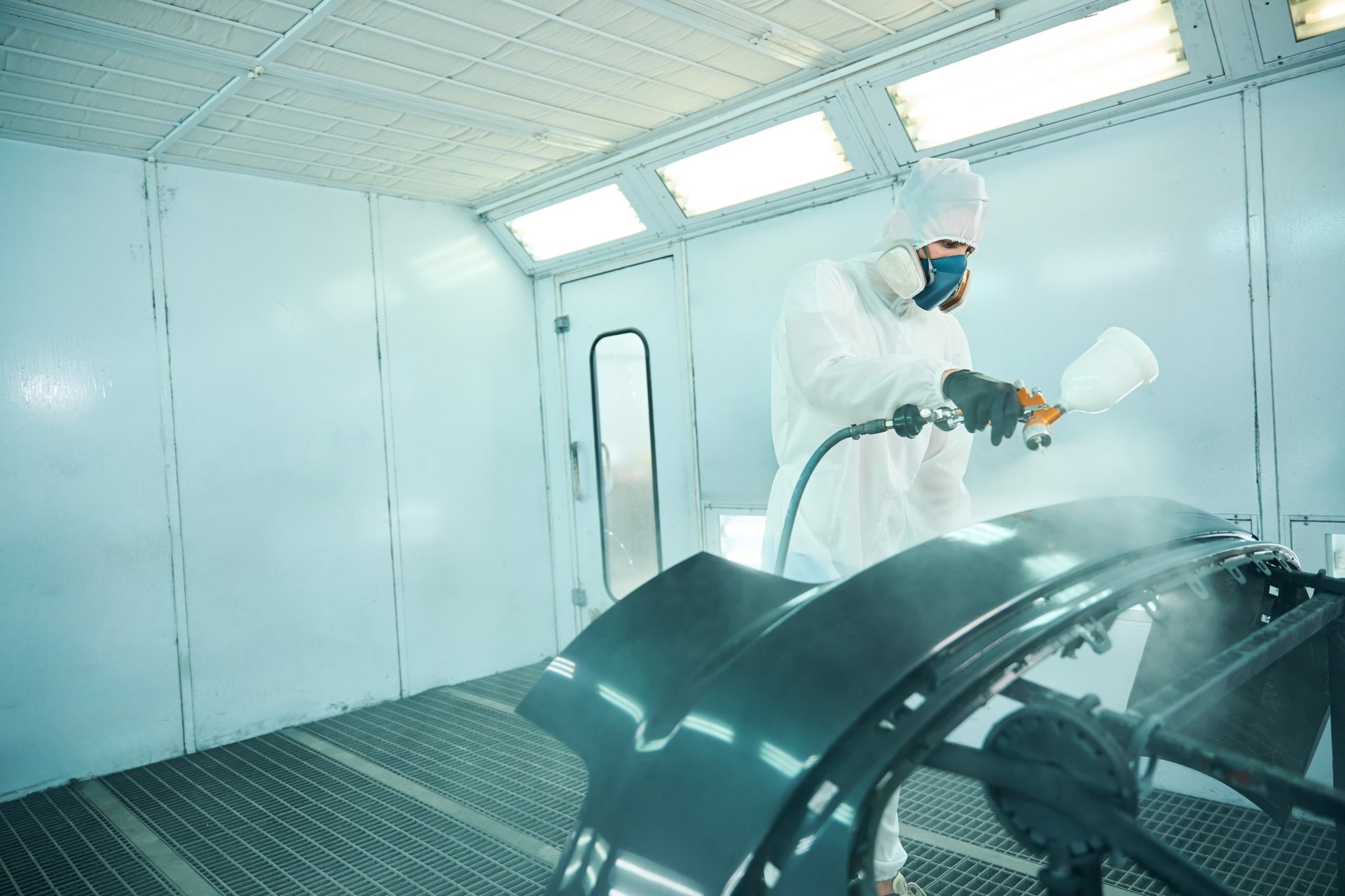 Mechanic in protective uniform painting car using spray compressor in workshop